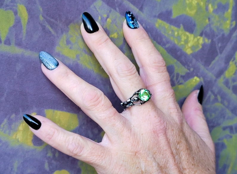 May include: A hand wearing a silver ring with a green gemstone. The ring has a skeletal design. The hand is also wearing black and silver nail polish.