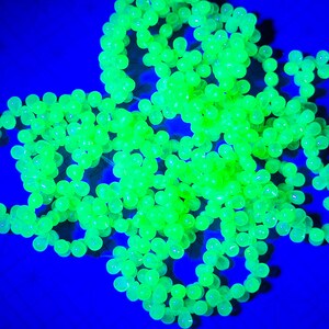 K&ouml;nnte beinhalten: Eine Ansammlung von leuchtend gr&uuml;nen Glasperlen, die auf einem blauen Hintergrund in einem Sternenausbruch angeordnet sind. Die Perlen sind durchscheinend und leuchten unter Schwarzlicht.