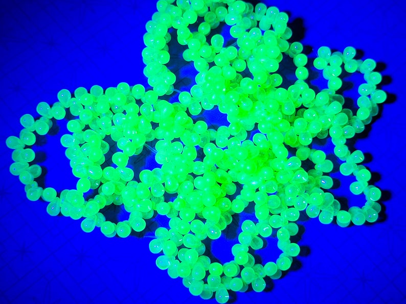 K&ouml;nnte beinhalten: Eine Ansammlung von leuchtend gr&uuml;nen Glasperlen, die auf einem blauen Hintergrund in einem Sternenausbruch angeordnet sind. Die Perlen sind durchscheinend und leuchten unter Schwarzlicht.