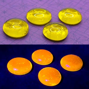 Puede incluir: Cuatro cabujones de vidrio amarillo, lisos y circulares, se muestran bajo dos condiciones de iluminación. La vista superior los muestra con luz natural, mientras que la inferior muestra los mismos cuatro cabujones brillando en naranja bajo luz UV.
