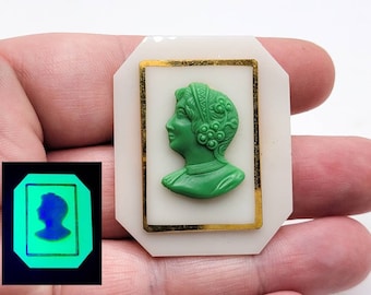 May include: A white cameo brooch with a gold frame. The cameo features a green profile of a woman with a floral headdress.