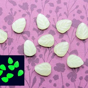 May include: Ten white leaf-shaped glow-in-the-dark beads on a pink floral background. The beads are arranged in a semi-circle. A smaller image shows the beads glowing green in the dark.