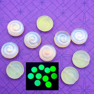 May include: A collection of iridescent, spiral-shaped beads and round, translucent beads. A separate image shows the round beads glowing bright green. The beads are arranged on a purple surface with a star pattern.