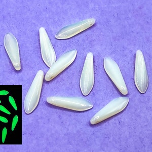 Puede incluir: Un grupo de diez cuentas blancas e iridiscentes en forma de lágrima. Las cuentas están dispuestas sobre una superficie morada. Una pequeña imagen cuadrada en la esquina inferior izquierda muestra las cuentas brillando en verde en la oscuridad.