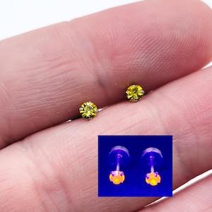 Puede incluir: Dos pequeños pendientes de botón con piedras preciosas amarillas. Los pendientes tienen un engaste de color plateado. Una imagen más pequeña muestra los pendientes brillando bajo luz negra.