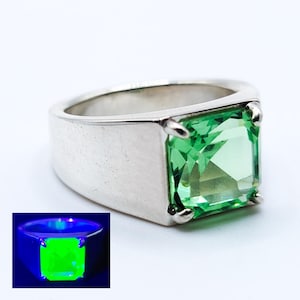May include: A silver ring featuring a large, square-cut, bright green gemstone. The ring has a thick band and is set with four prongs. A smaller image shows the gemstone glowing green under UV light.