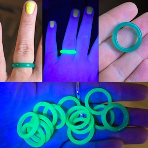 May include: A collection of green and glowing rings. One ring is on a finger, and others are held in a hand. The rings appear green in normal light and glow brightly under a black light. The rings are a solid color and have a smooth surface.