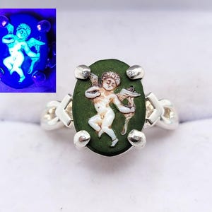 Può includere: Anello in argento con una pietra ovale verde scuro, raffigurante un cherubino con arco e freccia. L'anello ha un design distintivo, con quattro accenti argentati attorno alla pietra e una fascia a motivi. L'immagine mostra anche una versione luminosa del cherubino.