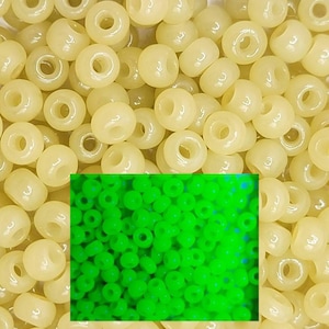 May include: A close-up image of a pile of small, round, opaque, pale yellow beads. The beads are arranged in a random pattern. A small square image in the lower right corner shows the same beads glowing green under a black light.