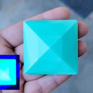May include: A turquoise square object with a pyramidal design is held in a hand. The object has a glossy finish and is illuminated by a light source. A smaller, glowing square is in the lower left corner.