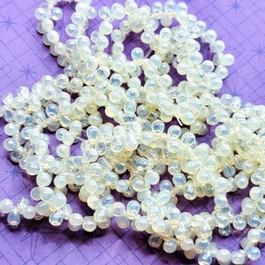 K&ouml;nnte beinhalten: Eine Str&auml;hne aus irisierenden wei&szlig;en Glasperlen, jede Perle hat die Form einer Tr&auml;ne. Die Perlen sind auf einem durchsichtigen Faden aufgef&auml;delt und auf einem violetten Hintergrund verstreut.