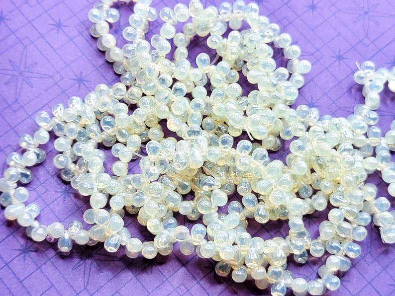 K&ouml;nnte beinhalten: Eine Str&auml;hne aus irisierenden wei&szlig;en Glasperlen, jede Perle hat die Form einer Tr&auml;ne. Die Perlen sind auf einem durchsichtigen Faden aufgef&auml;delt und auf einem violetten Hintergrund verstreut.