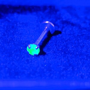 May include: A green gemstone set in a silver body piercing. The gemstone is glowing under a UV light.