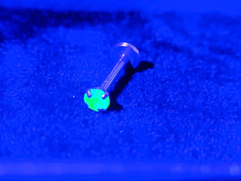 May include: A green gemstone set in a silver body piercing. The gemstone is glowing under a UV light.