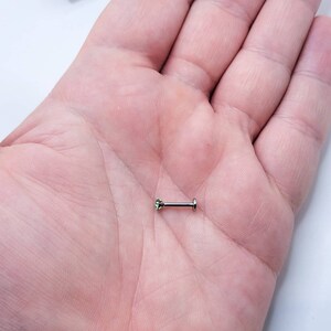 May include: A small, silver barbell with a green gemstone on one end. The barbell is lying on a person's palm.