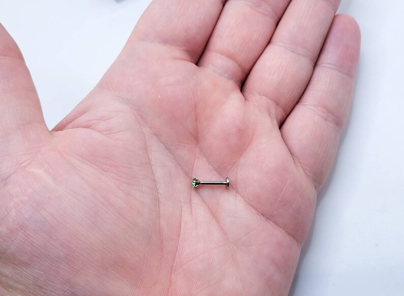 May include: A small, silver barbell with a green gemstone on one end. The barbell is lying on a person's palm.