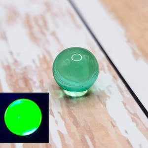 以下が含まれることがあります： 半透明で丸い緑色のガラス玉。大理石にはわずかな渦巻き模様があり、明るい色の木製の表面に置かれています。小さな正方形のインセットは、黒い背景に対して明るい緑色に輝く大理石を示しています。