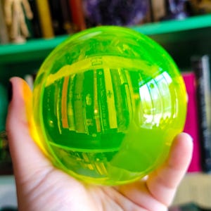 Puede incluir: Una esfera translúcida de color verde neón se sostiene en una mano. La esfera refleja una estantería de libros, creando una imagen distorsionada y colorida en su interior. La superficie de la esfera tiene un degradado de amarillo y verde.