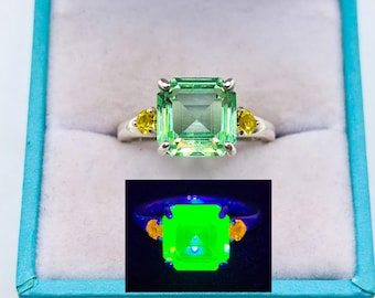 Könnte beinhalten: Ein Silberring mit einem großen, smaragdgeschliffenen hellgrünen Mittelstein, flankiert von zwei kleineren runden gelben Seitensteinen. Ein Inset zeigt denselben Ring unter UV-Licht, wobei der Mittelstein neongrün und die Seitensteine orange leuchten.