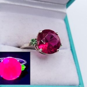 Puede incluir: Un anillo de plata con una gran gema magenta facetada y dos piedras verdes más pequeñas. El anillo se muestra en una caja de joyería azul claro, con una imagen más pequeña y brillante del anillo.