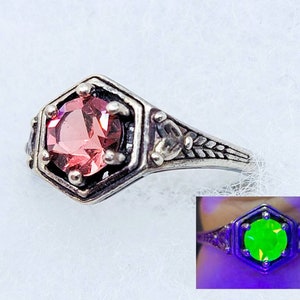 May include: A silver ring with a pink gemstone set in a hexagonal bezel. The ring has a delicate floral design on the band. The gemstone glows green under UV light.