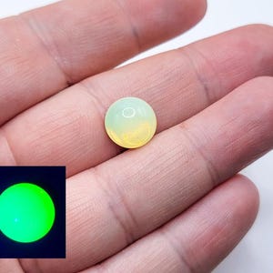 May include: A round, pale yellow and green gemstone, held in a hand. The stone has a gradient of color, transitioning from yellow to green. A small, square image in the lower left corner shows the stone glowing bright green.