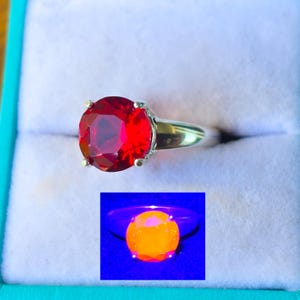 May include: A silver-toned ring featuring a large, round, faceted red gemstone held by four prongs. An inset shows the same ring under ultraviolet light, where the gemstone exhibits a bright, glowing orange fluorescence against a dark background.