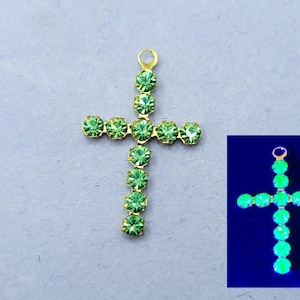 Puede incluir: Un colgante de cruz de oro con diamantes de imitación verdes. La cruz se muestra bajo una luz negra, revelando que los diamantes de imitación brillan en la oscuridad.