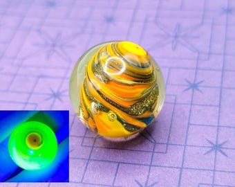 May include: A clear glass marble featuring a vibrant swirl of orange, yellow, and black. The marble has a glittery texture and is set against a purple backdrop. A smaller, glowing green and blue image is in the lower left corner.