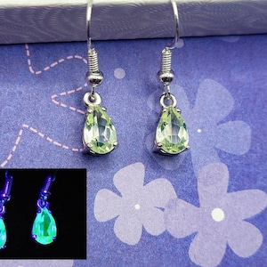May include: A pair of silver teardrop earrings with green stones that glow in the dark. The earrings are shown in a close-up image and a separate image showing the stones glowing.