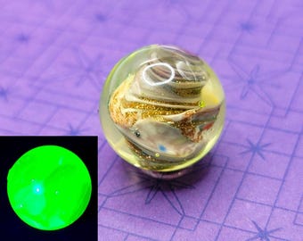 May include: A clear glass marble with swirling gold, white, and brown inclusions. The marble has a glittery appearance and is set against a purple background. A smaller, glowing green circle is in the lower left corner.