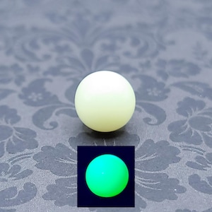 Puede incluir: Una bola redonda de color blanquecino, posiblemente una cuenta o elemento decorativo. Una imagen más pequeña insertada muestra la bola brillando en verde brillante sobre un fondo oscuro. La bola parece lisa y esférica, con un brillo suave y difuso.