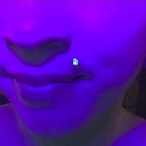 May include: A close-up of a person's face under a blacklight. The person has a small, green, glowing stud in their lower lip piercing. The person's ear is also pierced with a large, round, blacklight-reactive earring.