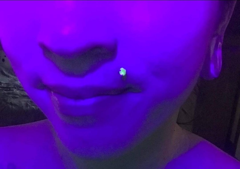 May include: A close-up of a person's face under a blacklight. The person has a small, green, glowing stud in their lower lip piercing. The person's ear is also pierced with a large, round, blacklight-reactive earring.