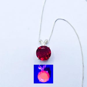 Puede incluir: Un collar de plata con un colgante de piedra preciosa redonda y facetada de color rojo intenso, sujeto por un engaste de garras de plata. Un recuadro muestra el mismo colgante brillando con una fluorescencia naranja neón bajo luz ultravioleta.