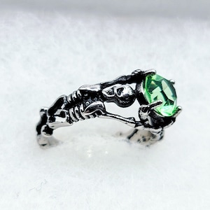 May include: A silver ring with a skeleton holding a large green gemstone.