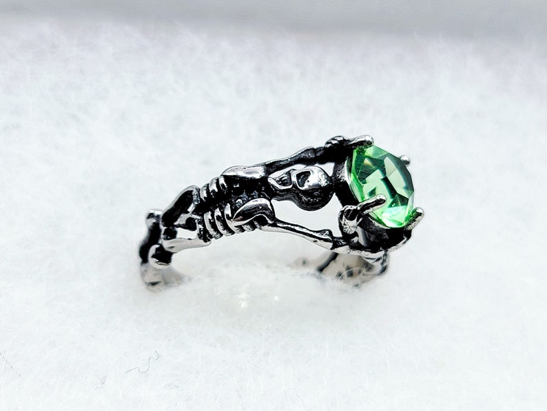 May include: A silver ring with a skeleton holding a large green gemstone.