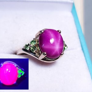 May include: A silver ring featuring a large, oval, purplish-pink gemstone with a star-like effect. The ring is accented with small green gemstones. The ring is displayed in a light blue jewelry box.