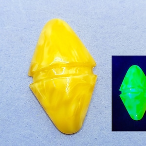May include: A yellow, diamond-shaped object with a rounded top and bottom. The object has a textured surface and a groove in the middle. A smaller image shows the object glowing green under a black light.