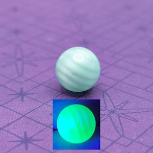 Puede incluir: Una canica de vidrio verde claro y blanco con remolinos. La canica es redonda y translúcida. Una imagen más pequeña muestra la canica brillando en verde brillante.