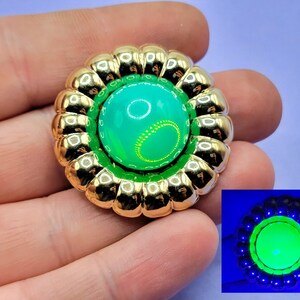 Puede incluir: Un broche de forma de flor de tono dorado con un cabujón de vidrio verde en el centro. El cabujón brilla en verde bajo una luz negra.