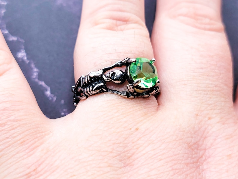 May include: A silver ring with a skeleton design holding a large green gemstone.