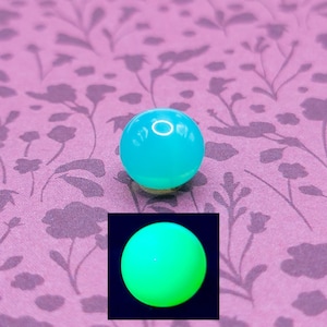 Puede incluir: Una esfera luminosa de color turquesa, posiblemente una cuenta o elemento decorativo, se muestra sobre un fondo morado estampado. Una imagen más pequeña insertada muestra la esfera brillando en verde brillante. La esfera parece estar hecha de un material translúcido.