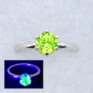 May include: A silver ring with a green gemstone that glows under a black light. The ring is set with a round, faceted stone.