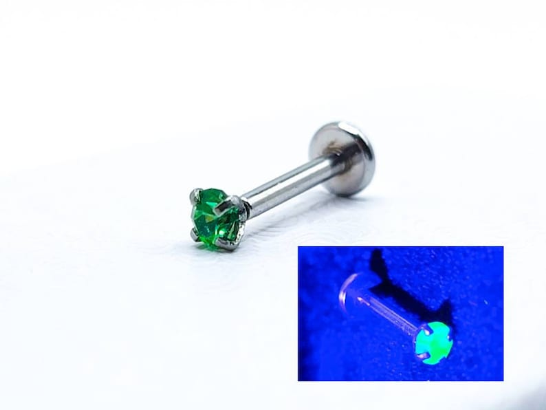 May include: A silver body piercing with a small green gem. The gem glows green under a black light.