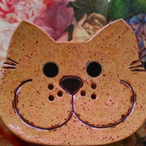 Op de afbeelding: Een keramische zeepbak in de vorm van een kat met een gespikkelde oranje glazuur. De zeepbak heeft een neus die omhoog steekt, twee zwarte ogen en een kleine mond met twee gaten aan weerszijden.