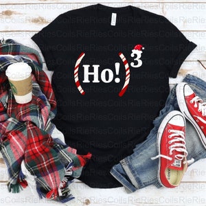 May include: Black t-shirt with a white candy cane design and the text "Ho! 3" with a Santa hat on the 3. The shirt is laid out on a wooden surface with a red and black plaid scarf, a pair of blue jeans, red sneakers, and sunglasses.