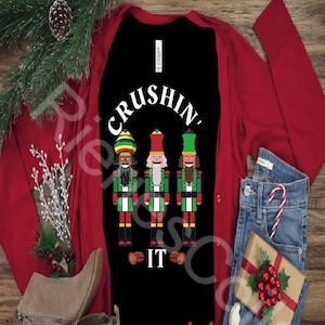 May include: A black long-sleeved shirt with the text "CRUSHIN' IT" in white. Three nutcracker figures are depicted below the text, each with a different skin tone and facial features. The shirt is worn over a red cardigan and paired with brown ankle boots.