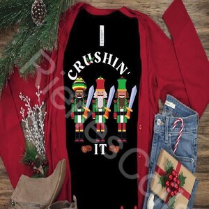 May include: A black long-sleeved shirt with the text "CRUSHIN' IT" in white. Three nutcracker figures are depicted in the center of the shirt, each with a different skin tone and a sword in hand. The shirt is worn over a red cardigan and paired with blue jeans.