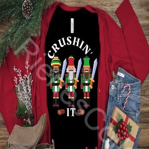 May include: A black long-sleeved shirt with a red cardigan and blue jeans. The shirt has a graphic of three nutcrackers with the text "CRUSHIN' IT".